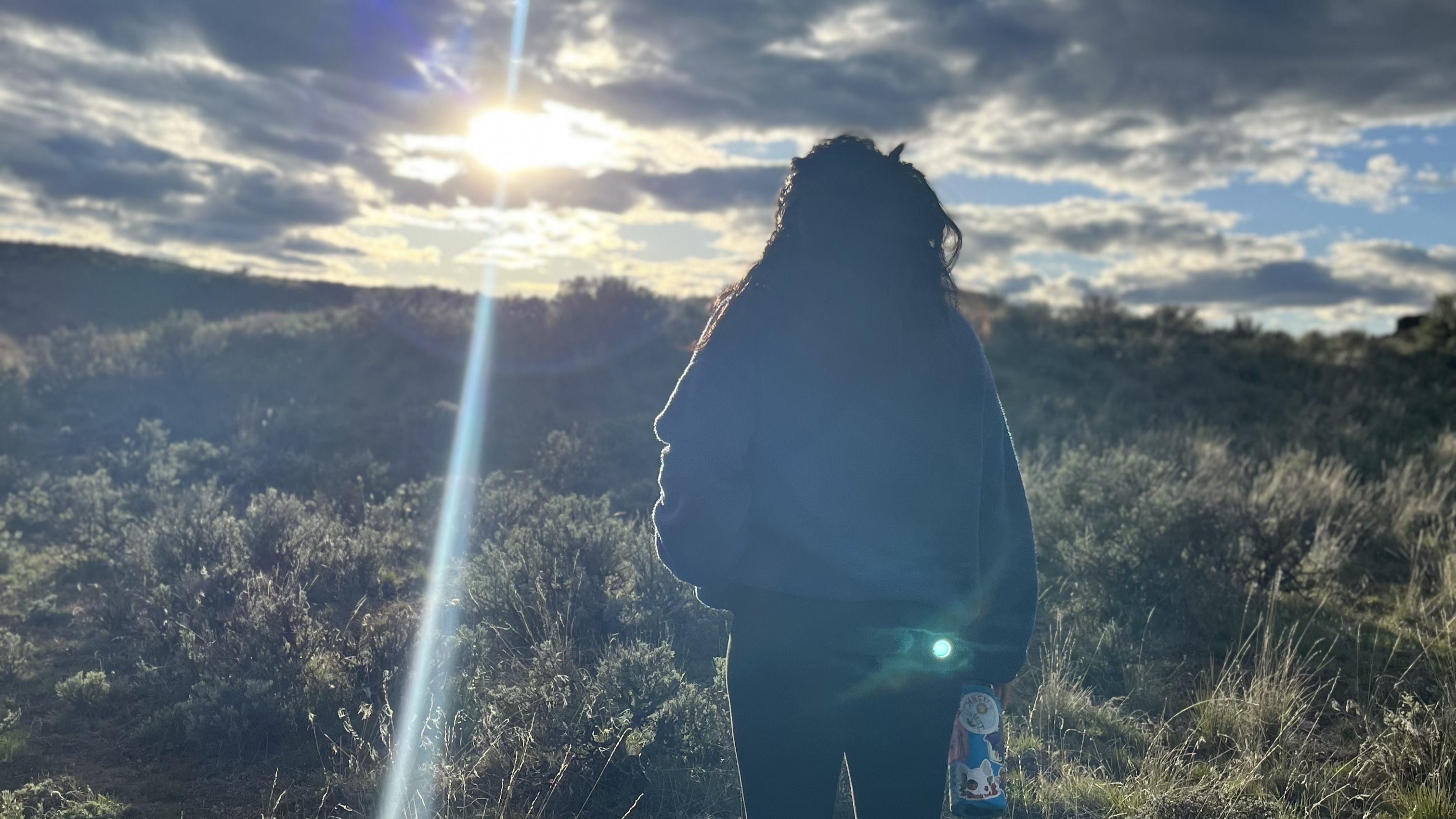 Maria’s silohette looking into the distance of a high desert with the sun in the clouded sky.