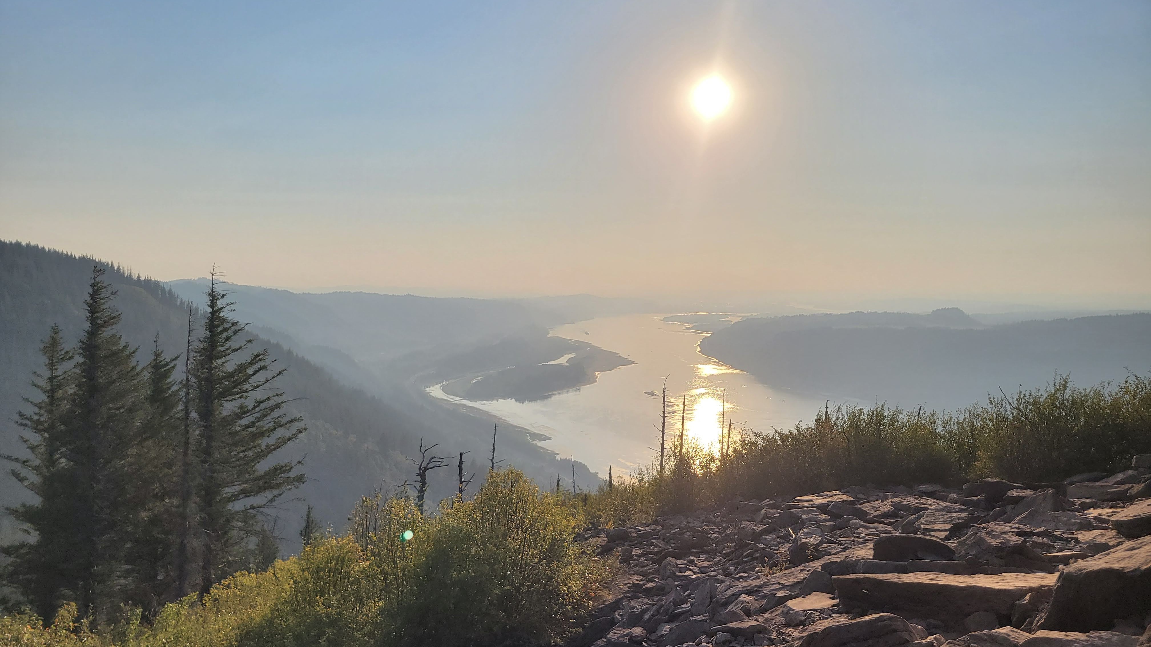 Forested mountainsides with a river flowing in the middle into the distance, with the sun in the sky.